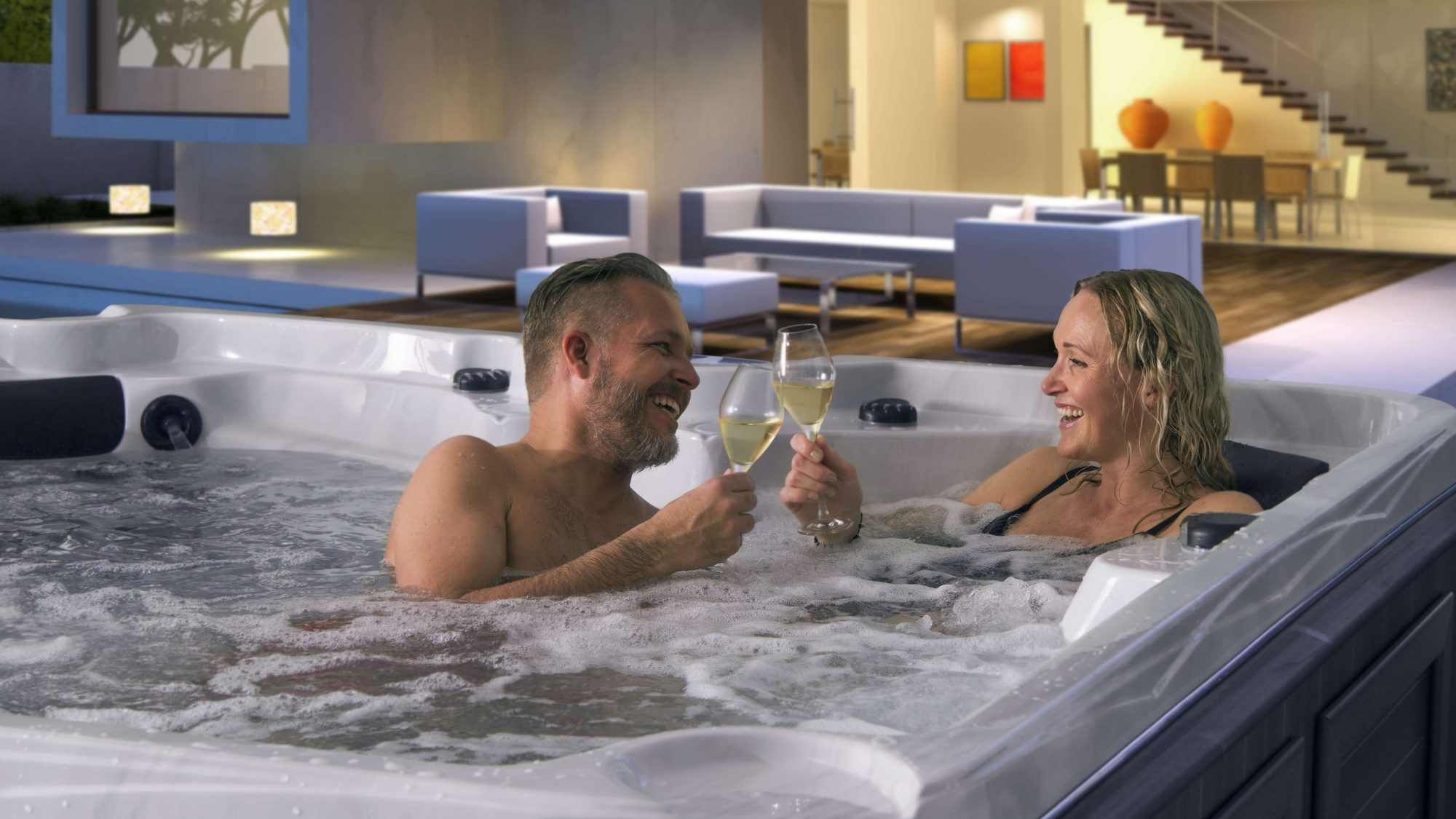 Un couple trinque au verre de vin dans un spa Arctic Spas, le soir, devant le salon éclairé d'une maison moderne